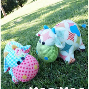 Peut inclure: Deux jouets en peluche en forme de vache, l'un rose et blanc à pois et l'autre vert et blanc à motifs d'étoiles. Les deux vaches ont des yeux noirs et un nez rose.