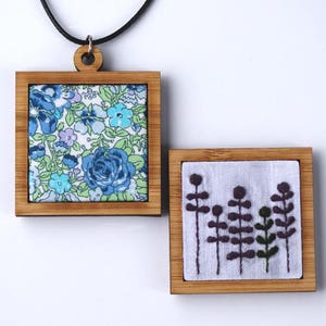 May include: Two square wooden frames with fabric inside. The top frame has a floral pattern in blue, green, and white. The bottom frame has a white fabric with embroidered purple flowers.