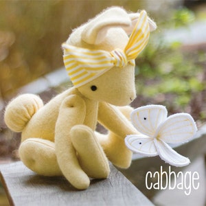 Könnte beinhalten: Ein weiches, gelbes Hasen-Plüschtier mit einem gestreiften gelb-weißen Stirnband. Der Hase hält ein weiß-gelbes, kohlförmiges Spielzeug. Das Wort "cabbage" steht in weißer Schrift.