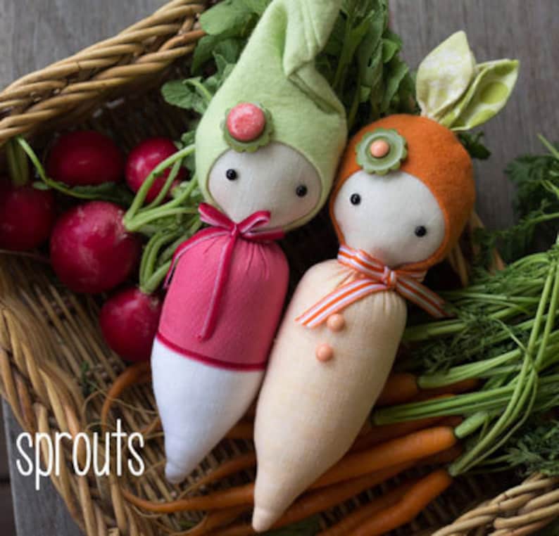 Pode incluir: Duas bonecas de tecido em forma de vegetais, um rabanete rosa e branco e uma cenoura laranja, com olhos de bot&atilde;o e chap&eacute;us decorativos. Est&atilde;o numa cesta de vime com rabanetes e cenouras. A palavra "sprouts" est&aacute; escrita em branco.