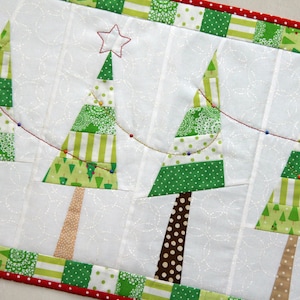 May include: A white fabric runner with a red and green border features a string of lights and four patchwork Christmas trees. The trees are made of green, white, and brown fabric with a variety of patterns.
