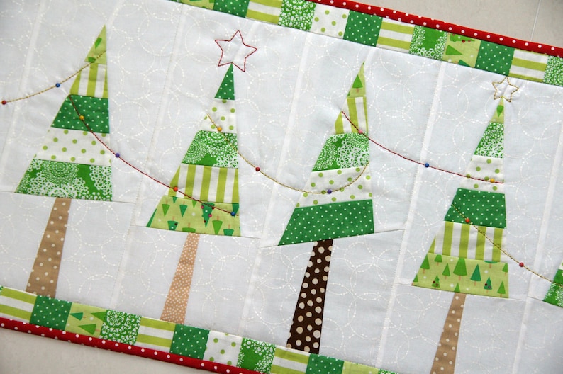 May include: A white fabric runner with a red and green border features a string of lights and four patchwork Christmas trees. The trees are made of green, white, and brown fabric with a variety of patterns.