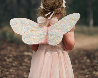 May include: A young child wearing a pink tulle dress and butterfly wings with a floral pattern. The wings are made of fabric and have a light blue, pink, and yellow color scheme.