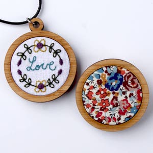 May include: Two round wooden framed embroidery hoops. One hoop features a white fabric with the word "Love" embroidered in teal blue with purple and yellow flowers. The other hoop features a colorful floral fabric with red, blue, and yellow flowers.