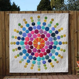Puede incluir: Una colcha colorida con un diseño circular que presenta una variedad de colores, incluyendo amarillo, naranja, rojo, rosa, morado, azul y verde. La colcha está hecha de círculos de diferentes tamaños, con el círculo más grande en el centro. La colcha está colgada en una valla de madera.