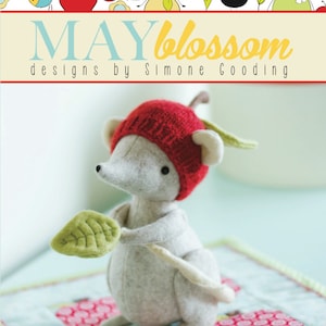 May include: A gray felt mouse wearing a red apple hat with a green felt leaf in its hand. The mouse is sitting on a blue and white patchwork quilt with red and white apple designs. The text "apple strudel the little shrew" is printed below the mouse.