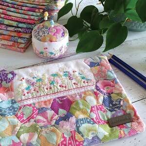 May include: A white fabric pouch with a floral design and a small embroidered flower border. The pouch is made of patchwork fabric with a variety of colors and patterns. The pouch has a brown leather tag with the word "handmade" printed on it.