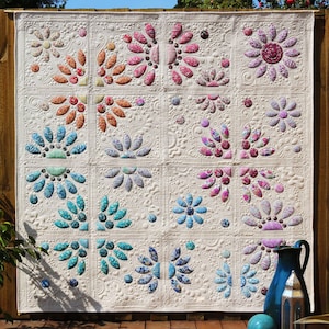 Op de afbeelding: Een wit quilt met een bloemenpatroon. Het quilt is gemaakt van 16 vierkanten, elk met een ander bloemenontwerp. Het quilt is gestikt met een reliëfpatroon, waardoor het een 3D-effect krijgt.