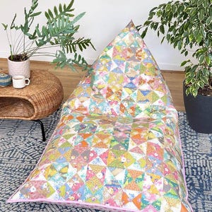 May include: A large, colourful patchwork floor cushion with a pink border. The cushion is made of many different fabrics, including floral prints, geometric patterns, and solid colours. The cushion is sitting on a blue and white patterned rug.