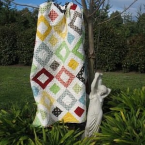 May include: A colorful patchwork quilt with a diamond pattern in shades of red, yellow, green, blue, and white. The quilt is hanging on a tree branch in a garden setting.