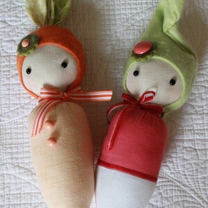 Pode incluir: Duas bonecas de tecido feitas &agrave; m&atilde;o. Uma boneca &eacute; laranja com orelhas de coelho verdes e um detalhe de flor. A outra boneca &eacute; vermelha e branca com um chap&eacute;u verde. Ambas as bonecas t&ecirc;m olhos pretos e fitas.