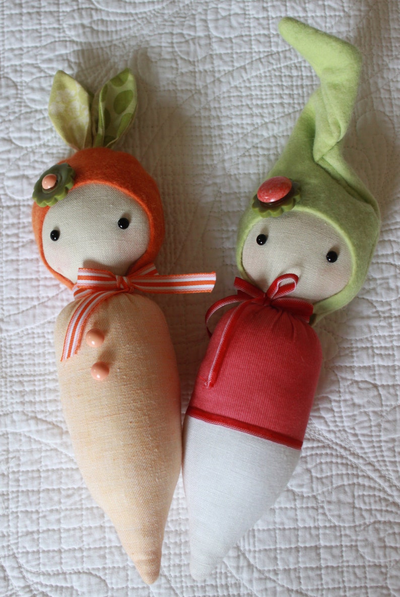 Pode incluir: Duas bonecas de tecido feitas &agrave; m&atilde;o. Uma boneca &eacute; laranja com orelhas de coelho verdes e um detalhe de flor. A outra boneca &eacute; vermelha e branca com um chap&eacute;u verde. Ambas as bonecas t&ecirc;m olhos pretos e fitas.