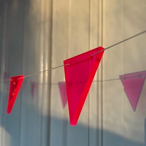 Könnte beinhalten: Eine Girlande aus rosa dreieckigen Flaggen, die an einer weißen Schnur hängen. Die Flaggen bestehen aus einem durchscheinenden Material und werden von Sonnenlicht beleuchtet.