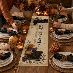 Pode incluir: Um caminho de mesa retangular longo com o texto 'Sophie's Coven' e 'Members Since 2026'. O design inclui ilustrações de um livro de feitiços, uma vela e folhagem de outono. Está centrado numa mesa de jantar de madeira com lugares postos.