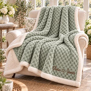 May include: A sage green throw blanket with a textured waffle-knit pattern and a cream-coloured sherpa fleece lining. The blanket is draped over an armchair and features the embroidered text Olivia and James with a heart symbol in brown thread.