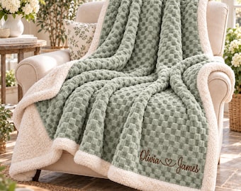 May include: A sage green throw blanket with a textured waffle-knit pattern and a cream-colored sherpa fleece lining. The blanket is draped over an armchair and features the embroidered text Olivia and James with a heart symbol in brown thread.
