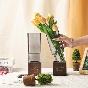 Puede incluir: Dos jarrones de cristal transparente, uno vertical y otro inclinado, cada uno sobre una base de madera. El jarrón inclinado contiene tulipanes amarillos. Las bases de madera están grabadas con iniciales y nombres. También se ven una pequeña planta en maceta y un marco de fotos.