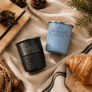 May include: Two metal tumblers with a textured lower section and a smooth upper rim. One is matte black with a cabin and tree graphic labelled The Miller Family, and the other is light blue with a mountain and tree graphic labelled The Anderson Family.