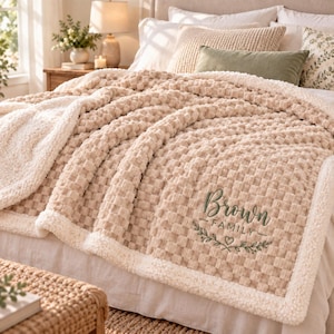 May include: A beige textured throw blanket with a white sherpa fleece border. The blanket features the embroidered text Brown FAMILY in dark green, accompanied by a small heart and leaf design, draped over a bed with pillows.