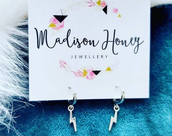 Pode incluir: Brincos de argola de prata com um charme de raio. Os brincos são exibidos em um cartão branco com o texto "Madison Honey Jewellery".