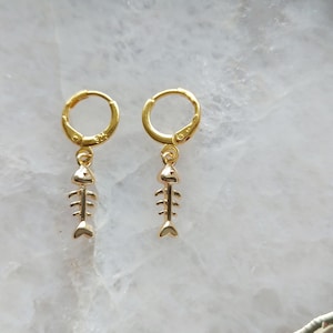 Puede incluir: Pendientes de aro dorados con un colgante de espina de pescado. Los pendientes son simples y delicados, perfectos para el uso diario.