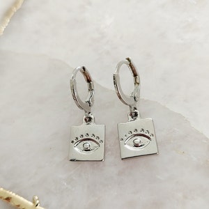 May include: A pair of silver hoop earrings with a square charm featuring an eye design. The eye is surrounded by a circle of small dots.