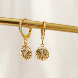 Puede incluir: Un par de pendientes de aro dorados con colgantes de concha. Los pendientes cuelgan de una barra dorada sobre un fondo blanco. Los colgantes de concha tienen crestas detalladas.