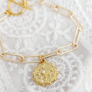 May include: A gold chain bracelet with a toggle clasp and a round pendant featuring an ornate design and a small white stone in the centre.