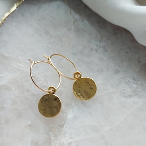 Könnte beinhalten: Ein Paar goldene Creolen mit einem kleinen, gehämmerten goldenen Scheibenanhänger, der an jedem Creolen hängt.