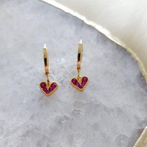 May include: A pair of gold hoop earrings with a dangling heart charm. The heart charm is decorated with small pink gemstones.