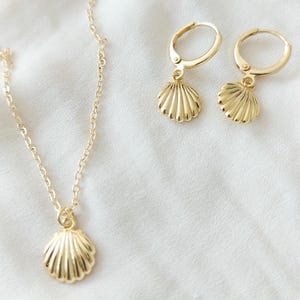 May include: A gold-coloured jewellery set featuring a seashell pendant necklace and matching seashell hoop earrings. The necklace has a delicate chain, and the earrings have a hinged closure. The jewellery is displayed on a white surface.