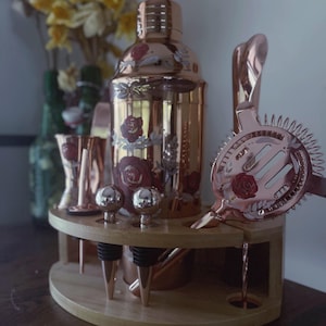 May include: A rose gold cocktail shaker set with a wooden stand. The set includes a shaker, jiggers, a strainer, bottle stoppers, and other bar tools. The shaker and strainer are decorated with red rose designs.