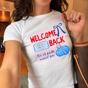 May include: A white short-sleeved t-shirt featuring a graphic with pink vertical stripes, a blue bow, and the red text "WELCOME BACK". Below, a blue rectangular cold pack and a blue round ice bag are depicted with the text "the ice packs missed you".