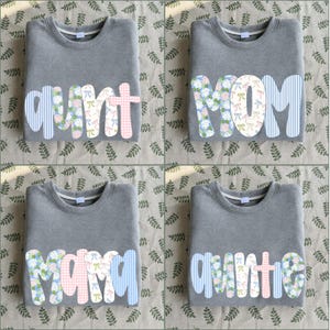May include: Four grey crewneck sweatshirts featuring large, fabric-appliqued letters on the front. The letters spell out "aunt", "MOM", "mama", and "auntie" using a mix of blue striped, floral, and pink gingham patterned fabric patches.
