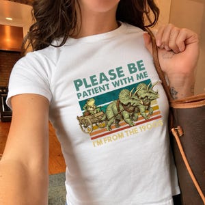 Op de afbeelding: Een wit t-shirt met korte mouwen en een afbeelding van een kikker in een houten kar getrokken door twee triceratops dinosaurussen. De tekst "PLEASE BE PATIENT WITH ME" en "I'M FROM THE 1900S" staat boven horizontale gekleurde strepen.