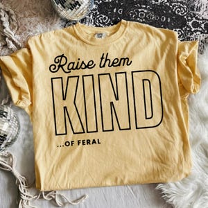 May include: A light yellow t-shirt with the words "Raise them KIND ...OF FERAL" in black lettering. The shirt is made of a soft material and has a crew neck.
