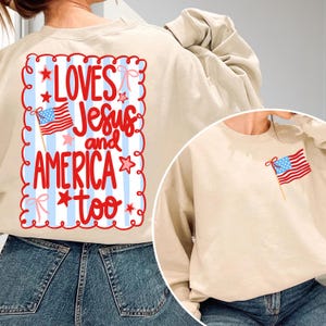 May include: A beige crewneck sweatshirt featuring a back graphic with a light blue striped background, a red scalloped border, and red text that reads LOVES Jesus and AMERICA too, accented with stars and bows. The front features a small American flag.