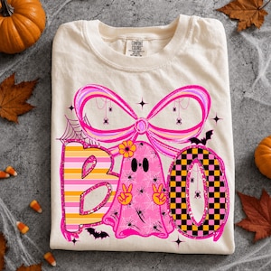 Może przedstawiać: Kremowy t-shirt z grafiką różowego duszka z kwiatkiem, wykonującego znak pokoju. Wzór zawiera dużą różową kokardę, pajęczyny, małe pająki, nietoperze oraz napis BOO w paski i szachownicę.