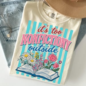 May include: A cream-colored t-shirt featuring a graphic of an open book with colorful flowers emerging from the pages. The text reads "it's too NONFICTIONY outside" in red, pink, and blue. The background design consists of vertical light blue stripes.