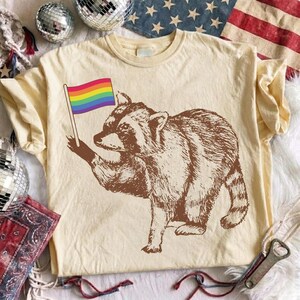 Puede incluir: Camiseta color crema con una ilustración de un mapache marrón sosteniendo una bandera arcoíris. La ilustración del mapache es detallada, con los colores de la bandera vibrantes contra el tono neutro de la camiseta. La camiseta tiene un diseño de cuello redondo clásico.
