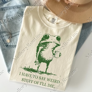 Op de afbeelding: Crèmekleurig T-shirt met een groene kikkerprint die een mok vasthoudt. De tekst "I HAVE TO SAY WEIRD STUFF OF I'LL DIE" staat eronder. Een bruine hoed en een blauwe jeans zijn op de achtergrond.