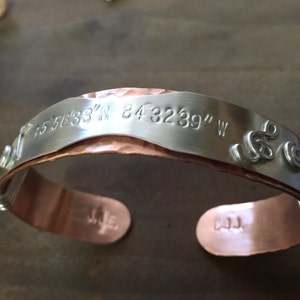 May include: A copper and silver bracelet with a hammered copper band and a silver band engraved with the coordinates "35.5633° N 84.3239° W".