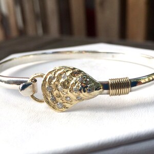 May include: A silver bangle bracelet with a gold clasp shaped like a seashell with small diamonds. The clasp is secured with a gold wire wrap.