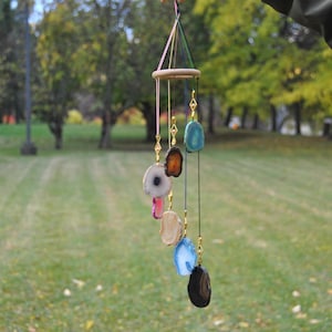 May include: A handmade wind chime with a wooden ring and colourful agate slices. The agate stones are in various colours, including blue, brown, and white, suspended by gold-coloured chains. The wind chime is hanging outdoors with a green background.