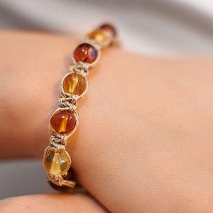 以下が含まれることがあります： 黄色と茶色のアンバービーズで作られたブレスレット。ビーズはベージュのコードに糸に通され、結び目で結ばれています。