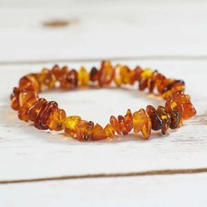 May include: A close-up of a brown amber bracelet made of small, irregular shaped beads. The bracelet is lying on a white wooden surface.