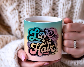 Może przedstawiać: Biały ceramiczny kubek z napisem "Love is in the Hair" w stylu retro. Kubek ma gradientowe tło w kolorach niebieskim, żółtym i różowym. Tekst otoczony jest chmurowym wzorem.