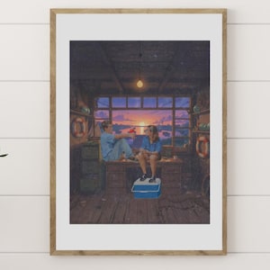 May include: A framed print depicting two people sitting inside a wooden structure, with a sunset visible through a window. The artwork is displayed on a white wall, with a wooden table and a decorative vase in the foreground.