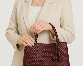 May include: A burgundy woven handbag with braided handles and tassel details. The rectangular bag is held by a person wearing a beige blazer and a white sweater. The bag's texture is visible, showcasing the intricate weave pattern.
