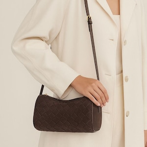 May include: A dark brown shoulder bag with a woven pattern. The bag has a long, thin strap and a gold-colored zipper. The bag is being held by a person wearing a cream-colored blazer.